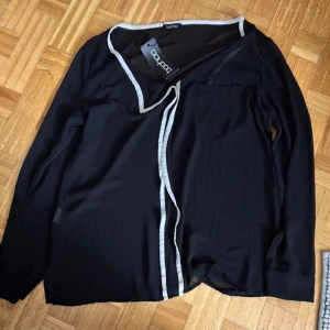 Svart omlottkofta från Boohoo - Svart omlottkofta från Boohoo med vita kontrastkanter och lång ärm. Koftan har en bröstficka och knytband framtill för en snygg och avslappnad look. Perfekt att slänga på sig över en topp eller klänning.