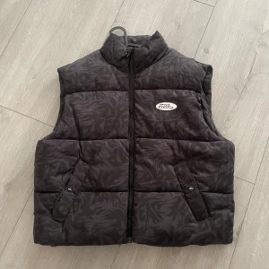 Svart dunväst från No Fear, mönstrad - Säljer en svart dunväst från No Fear i storlek M. Västen har ett diskret mörkt mönster, hög krage och dragkedja framtill. Två fickor med knappstängning och en oval No Fear-logga på bröstet. Perfekt för lager-på-lager och streetwear.