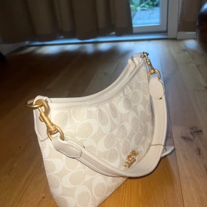 Beige axelväska från Coach - Snygg beige axelväska från Coach med klassiskt monogrammönster och guldfärgade metalldetaljer. Väskan har både kort och lång rem, dragkedja upptill och logga framtill. Perfekt storlek för det viktigaste och ger en clean och lyxig vibe.