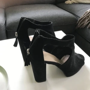 Svarta mockasandaler med klack - Snygga svarta sandaler i mocka med öppen tå och kraftig klack. Skorna har dragkedja bak för enkel påtagning och en cool, modern design med utskurna detaljer på sidorna. Perfekta för dig som vill ha en stilren men edgy look.