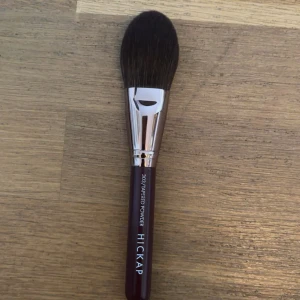 Hickap 303 Tapered Powder borste - Säljer en Hickap 303 Tapered Powder sminkborste med mörkbrunt borst, silverfärgat fäste och mörklila handtag. Perfekt för applicering av puder, bronzer eller rouge. Borsten är av syntetiskt material och har en avsmalnande form för exakt applicering.