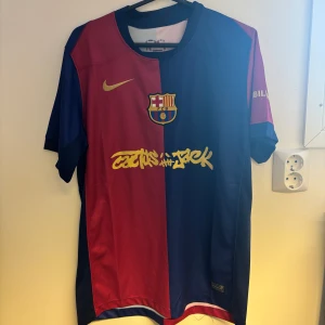 FC Barcelona x Cactus Jack matchtröja - Säljer en FC Barcelona x Cactus Jack matchtröja från Nike. Tröjan har klassisk halv-röd, halv-blå design med klubbmärke och gul Nike-logga på bröstet. Cactus Jack-graffiti och UNHCR-tryck i gult. Kortärmad och tillverkad i lätt, ventilerande material.