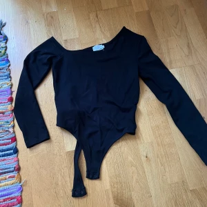 Svart långärmad bodysuit från Chiquelle - Säljer en svart långärmad bodysuit från NA-KD med offshoulder-skärning och markerade sömmar framtill och en öppen rygg. Bodyn har tryckknappar i grenen och är i stretchigt material som sitter tight mot kroppen. Perfekt för att skapa en clean och stilren look.
