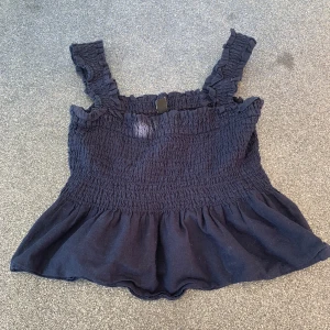 Mörkblå smockad linnetopp med volang - Supersöt mörkblå linnetopp med smockad överdel och breda axelband med volangdetaljer. Toppen har en utställd volang nertill som ger en lekfull look. Perfekt för varma dagar och enkel att styla med jeans eller kjol.