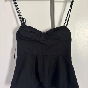 Svart peplum linnetopp med tunna band - Supersnygg svart linnetopp från Zara  med peplumvolang och tunna axelband. Toppen har söt rynkning vid bysten och smockad rygg för extra komfort. Perfekt att styla med jeans eller kjol för en trendig look.