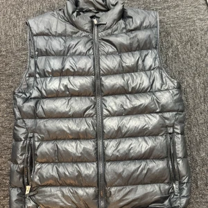 Svart dunväst från Gant - Snygg svart dunväst från Gant med quiltad design och hög krage. Västen har två fickor med dragkedja och stängs med en kraftig dragkedja framtill. Perfekt att slänga över en hoodie för en cool look.