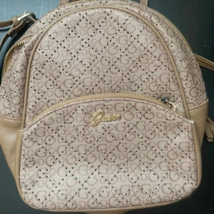 Beige ryggsäck Guess Manhattan City - Snygg beige ryggsäck från Guess, modell Manhattan City 34 cm. Väskan har all-over logomönster, guldfärgade detaljer och dragkedja. Materialet är syntet med skinnliknande känsla och insidan har Guess-logga. Perfekt storlek för vardagens essentials.