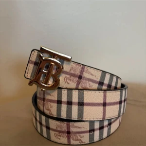 Rutig Burberry bälte med B-spänne - Snyggt bälte från Burberry med klassiskt rutigt mönster i beige, vinrött och grått. Bältet har ett stort metallspänne i guld med ett B framtill. Perfekt accessoar för att lyfta din outfit med en lyxig touch.