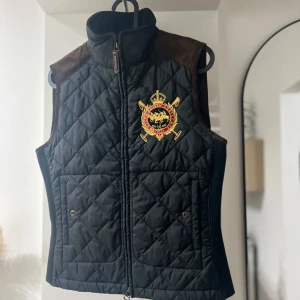 Svart väst Polo Ralph Lauren - Snygg svart quiltad väst från Polo Ralph Lauren med broderat emblem på bröstet och bruna mockadetaljer på axlarna. Västen har hög krage, dragkedja framtill och två fickor med knapp. 