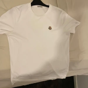 Vit Moncler t-shirt med logga - Snygg vit t-shirt från Moncler med klassisk rund hals och liten broderad logga på bröstet. Enkel och stilren design som passar perfekt till jeans eller shorts. Tillverkad i mjuk bomull för skön känsla hela dagen.