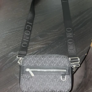 Svart axelväska från ICONO - Snygg svart axelväska från ICONO med justerbart band där loggan är broderad. Väskan har ett diskret mönster med bokstäver, silverfärgad dragkedja framtill och en metallring för extra detaljer. Perfekt storlek för mobilen och det viktigaste.