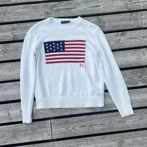 Vit stickad tröja Polo Ralph Lauren - Vit stickad tröja från Polo Ralph Lauren med USA flagga och RL-brodyr framtill. Klassisk rund halsringning, ribbade muddar och lång ärm. Perfekt för dig som gillar preppy stil och vill sticka ut med en ikonisk detalj.