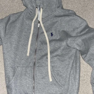 Grå hoodie från Polo Ralph Lauren - Snygg grå hoodie från Polo Ralph Lauren med dragkedja och vit snörning. Klassisk blå logga broderad på bröstet och stora fickor framtill. Mjuk insida och huva gör den perfekt för chill dagar. Passar dig som gillar stilren och sportig look.