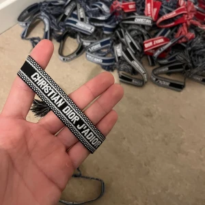Christian Dior J'Adior svart armband - Svart och vitt vävt armband från Christian Dior med texten 'CHRISTIAN DIOR J'ADIOR'. Armbandet har snygga tofsar i ändarna och ett justerbart knytband. Perfekt accessoar för att lyfta din outfit med en lyxig touch.