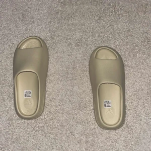 Beige Adidas Yeezy Slide - Adidas Yeezy Slide i beige med clean, minimalistisk design och öppen tå. Tillverkad i syntetmaterial med en chunky, räfflad sula som ger en modern vibe. Perfekt för chill dagar och enkel att matcha med streetwear.