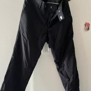 Svarta kostymbyxor med raka ben - Snygga svarta kostymbyxor med raka ben och klassisk passform. Byxorna har bälteshällor, två fickor fram och två bakfickor med knapp. Perfekta till en stilren look och passar till skjorta eller t-shirt. Nyskick, kostar närmare 1300:- ny.