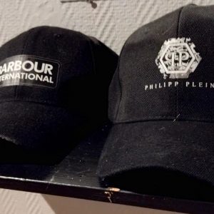 Svarta kepsar Barbour & Philipp Plein - Två svarta kepsar, en från Barbour International med vit logotyp framtill och en från Philipp Plein med vit PP-logga och text. Båda har klassisk böjd skärm och justerbar passform. Perfekta för en stilren cool och snygg outfit!
