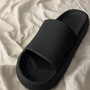 Svarta chunky slides i syntet - Svarta slides med chunky sula och ribbad ovandel. Skorna har öppen tå och är tillverkade i syntetmaterial, perfekta för chill dagar eller att glida runt hemma. Sulan har mönster och dräneringshål undertill för extra komfort.