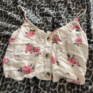 Blommig vit linnetopp med knappar - Supersöt vit linnetopp med rosa blommor och smala axelband. Toppen har tre knappar framtill och en croppad passform som ger en avslappnad och somrig vibe. Perfekt för varma dagar och lätt att matcha med jeans eller kjol. Är storlek 40 men är som en s/m