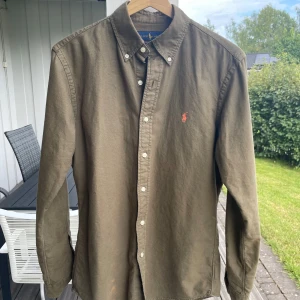 Olivgrön skjorta  Ralph Lauren - Olivgrön skjorta från Ralph Lauren med klassisk button down-krage och broderad logga i orange på bröstet. Skjortan är i bomull, har lång ärm och vita knappar framtill. Custom fit-modell. Mindre fläckar på ena sidan och ena ärmen men de är svaga