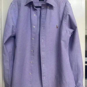 Lila skjorta från Ralph Lauren slim fit - Snygg lila skjorta från Ralph Lauren i slim fit-modell. Klassisk oxfordväv med knappar framtill och broderad logga på bröstet. Skjortan har lång ärm och är tillverkad i bomull med stretch för extra komfort.
