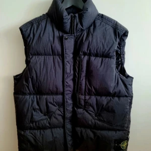 Svart dunväst från Stone Island - Säljer en svart dunväst från Stone Island med hög krage och dragkedja framtill. Västen har två stora fickor med knappstängning och den klassiska Stone Island-loggan på vänster sida. Tillverkad i nylon och fylld med dun för extra värme.