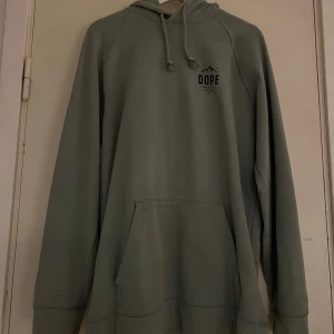 Grå hoodie från Dope med tryck - Snygg grå hoodie från Dope med svart tryck på bröstet och stort print på ryggen. Tröjan har huva med snörning och en stor magficka. 