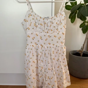 Vit blommig playsuit Hollister - Supersöt vit playsuit från Hollister med gula blommor, smala axelband och öppen rygg med resår. Klänningen har en luftig passform och är perfekt för varma dagar. 