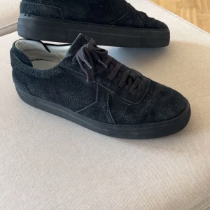 Svarta mockasneakers från Axel Arigato - Snygga svarta sneakers från Axel Arigato i mocka med svart platt sula och snörning. Skorna har en stilren design och de är perfekta för dig som gillar en clean look med premiumkänsla.