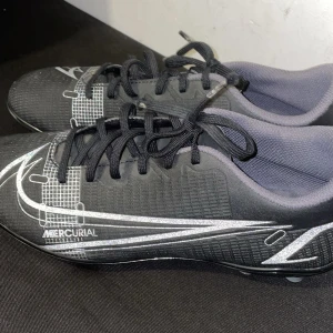 Nike Mercurial Fotbollsskor - Svarta Nike Mercurial fotbollsskor med silvriga detaljer och grafiskt mönster. Skorna har klassisk snörning som gör det enkelt att justera passformen efter foten. De är endast använda ett fåtal gånger och är därför i mycket gott skick. Tyvärr passade de inte, så de har mest legat oanvända i förrådet. Perfekta för dig som vill ha ett par snygga och välbevarade fotbollsskor från Nike till ett bra pris. 