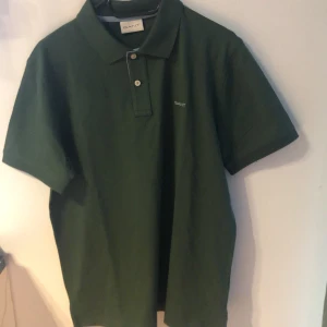 Grön pikétröja från GANT, storlek M - Snygg mörkgrön pikétröja från GANT i klassisk modell med korta ärmar och krage. Tröjan har två knappar framtill och diskret GANT-logga broderad på bröstet. Tillverkad i bomull för en skön och luftig känsla. Perfekt till jeans eller shorts.