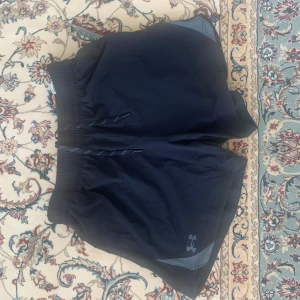 Svarta träningsshorts från Under Armour - Snygga svarta träningsshorts från Under Armour med elastisk midja och snörning. Shortsen har en diskret logga på benet och är gjorda i ett lätt och snabbtorkande syntetmaterial. Perfekta för gymmet eller löprundan.