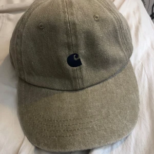 Beige Carhartt keps i bomull - Snygg beige keps från Carhartt med klassisk böjd skärm och mörkblå broderad logga framtill. Tillverkad i 100% bomull för en skön och avslappnad look. 