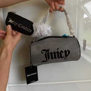 Juicy Couture glittrig handväska, cylindrisk  - Glittrig handväska från Juicy Couture i silver med svarta detaljer och logga. Väskan har cylinderform, dragkedja och matchande liten plånbok. Snygg metallkedja och blingiga stenar över hela väskan. Perfekt accessoar för att sticka ut.