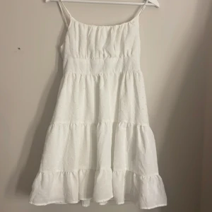 Vit somrig klänning med volanger - Superfin vit klänning med smala axelband och volanger i flera lager. Klänningen har broderade små detaljer i tyget och en smockad rygg för extra komfort. Perfekt för varma dagar och har en luftig, flowy känsla.