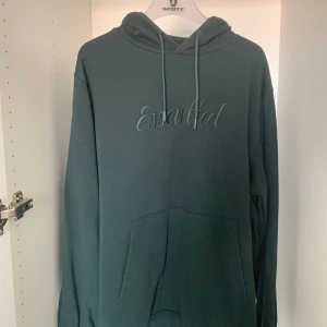 Mörkgrön hoodie från Dressmann Essential - Mörkgrön hoodie från Dressmann med broderad text 'Essential' på bröstet. Klassisk känguruficka framtill, huva med snörning och långa ärmar. Perfekt för dig som gillar en enkel och stilren look. Mjuk insida i bomullsblandning.