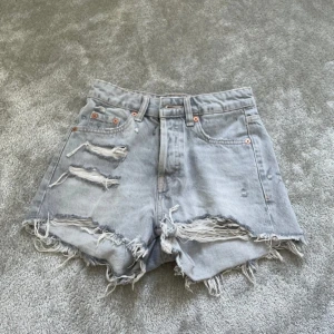 Gråa slitna jeansshorts från Zara - Snygga ljusgrå jeansshorts från Zara med hög midja och råa, fransiga kanter. Shortsen har flera slitningar och trasiga detaljer framtill för en cool, avslappnad look. Klassisk femficksmodell med knappgylf och bälteshällor.