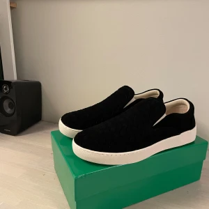 Svarta slip-on sneakers från Bottega Veneta - Stilrena svarta slip-on sneakers från Bottega Veneta med flätad ovandel i mocka och krämvit platt sula. Skorna har en minimalistisk look och är perfekta för dig som gillar lyxiga detaljer och enkel design. Skorna/loafersarna levereras med original boxen och Dustbags.
