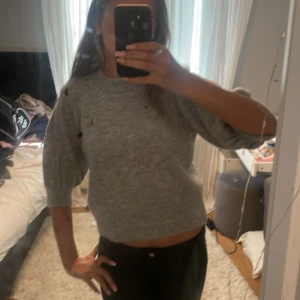 Grå stickad croppad tröja - Supermjuk grå stickad tröja med croppad passform och trekvartsärmar. Perfekt till höst och vinter, enkel att matcha med både jeans och kjol. Tröjan har en klassisk rund halsringning och ribbade muddar vid ärmslut och nederkant.