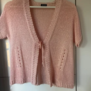 Ljusrosa stickad kofta från Gerry Weber - Supersöt ljusrosa stickad kofta från Gerry Weber med korta ärmar och djup v-ringning. Koftan har knytning framtill och två dekorativa fickor. Perfekt för lager-på-lager och ger en chill vibe till din outfit.