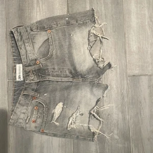 Grå slitna jeansshorts från Zara - Riktigt snygga grå jeansshorts från Zara med råa, fransiga kanter och slitningar framtill. Klassisk femficksmodell med knappar i gylfen och coolt, slitet uttryck. msydd för en mer trending och unik look. Billigt, snyggt och miljövänligt! Storlek 32