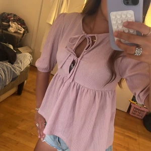 Ljusrosa blus med knyt och puffärm - Supersöt ljusrosa blus med knyt framtill och lätt puffade ärmar. Modellen är lite lös och har en flowy passform med rynk under bysten. Materialet är ribbat och toppen har en feminin, romantisk vibe. Perfekt till jeansshorts eller kjol för en chill look.