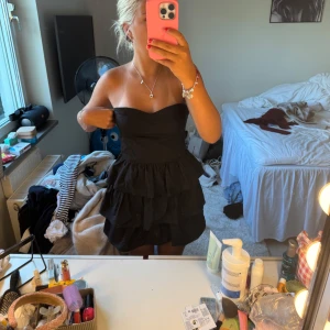 Svart axelbandslös volangklänning - Supersöt svart kort klänning med flera volanger och axelbandslös design. Sitter tajt upptill och har en fluffig kjol med lager på lager. Perfekt för dig som vill sticka ut med en klassisk men ändå lekfull look.