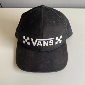 Svart keps från Vans med brodyr - Svart keps från Vans med klassisk böjd skärm och justerbar snapback. Framsidan har broderad Vans-logga i vitt med schackrutiga detaljer. Tillverkad i bomull med polyesterfoder. Perfekt för en streetwear-look.