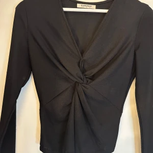 Svart omlottopp från Glamorous - Säljer en svart långärmad omlottopp från Glamorous med snygg twistad detalj framtill och djup v-ringning. Toppen har en tight passform och är perfekt för dig som gillar stilrena och trendiga plagg. Superenkel att matcha med både jeans och kjol.