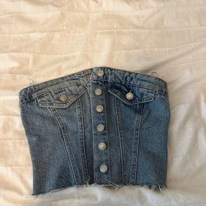 Jeanskorsett från ZARA med knappar - Cool jeanskorsett från ZARA i blå denim med silverfärgade knappar framtill och rå, fransig nederkant. Toppen har två dekorativa fickor och smock baktill för extra komfort. Perfekt att styla med höga byxor eller kjol.