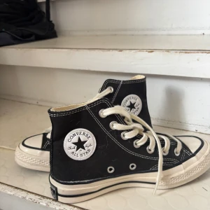 Svarta Converse Chuck Taylor All Star - Klassiska svarta Converse Chuck Taylor All Star med högt skaft och vita snören. Skorna har vit sula, rund tå och tydliga kontrastsömmar. Ikonisk logga på sidan och tillverkade i canvas för en tidlös streetstil.