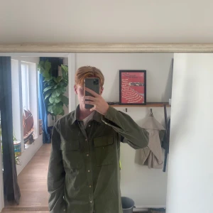 Grön Overshirt från East West - Säljer en grön overshirt från East West i regular fit. Overshirten har två bröstfickor med lock och knappar framtill. Färgen är två nyanser av grönt och materialet känns som bomull. Perfekt för dig som gillar en avslappnad stil.