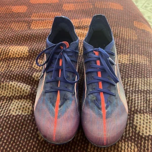 Blå och lila Puma fotbollsskor - Säljer ett par snygga Puma fotbollsskor i blått med rosa och vita detaljer. Skorna har snörning och mönstrad ovandel i syntetmaterial. Fotbollsskorna har en liten skada på sidan av skorna men inget som påverkar funktionen och spelet på plan.