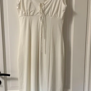 Vit långklänning från Gina Tricot - Superfin vit långklänning från Gina Tricot med snörning vid bysten och spetsdetaljer längs halsringningen. Klänningen har ärmlös design och mjukt, följsamt material som ger en snygg siluett.